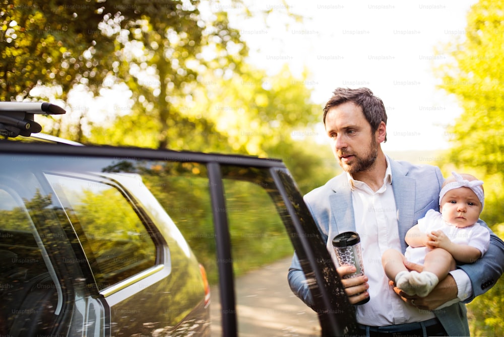 Junger Vater, der sein kleines Mädchen trägt. Mann am Auto, Kaffee zum Mitnehmen in der Hand.