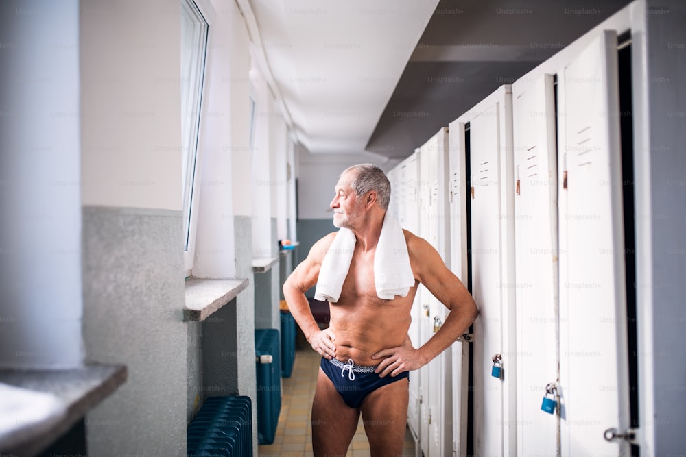 Senior man standing by the lockers in an indoor swimming pool, arms on hips. Active pensioner enjoying sport.