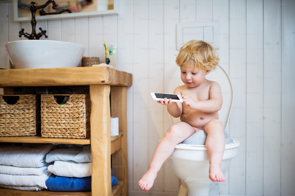 Süßes Kleinkind mit einem Smartphone im Badezimmer. Kleiner Junge, der auf der Toilette sitzt.
