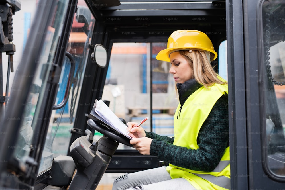 Female forklift truck driver in an industrial area. A woman sitting in the fork lift outside a warehouse, making notes.