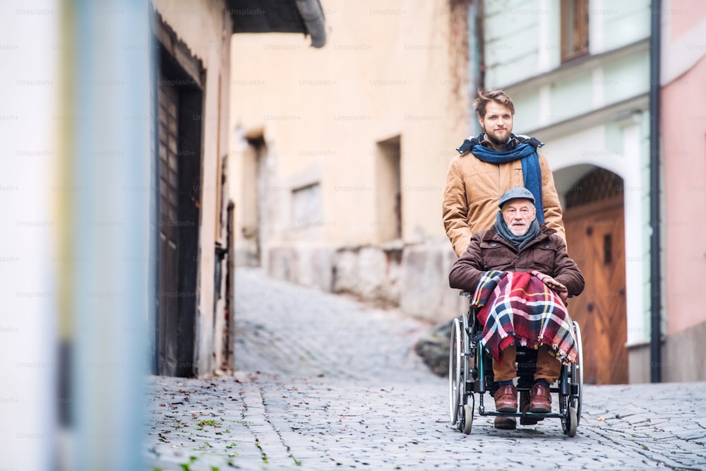 Père en fauteuil roulant et jeune fils en promenade. Un soignant s’occupe d’un homme âgé handicapé.