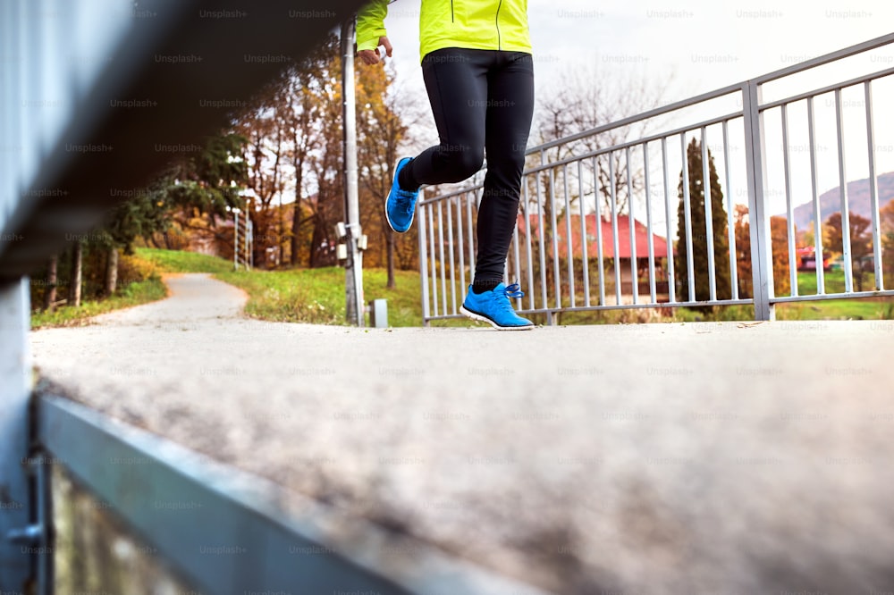 Unrecognizable young athlete in yellow jacket running outside by the lake. Trail runner training for cross country running in colorful sunny autumn nature.