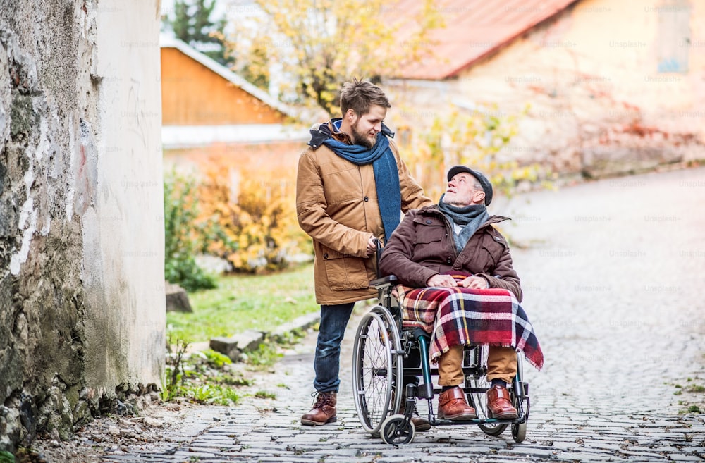 Père en fauteuil roulant et jeune fils en promenade. Un soignant s’occupe d’un homme âgé handicapé.