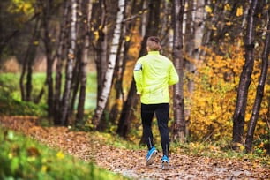 Young athlete with smartphone in yellow jacket running outside.Trail runner training for cross country running in colorful sunny autumn nature.