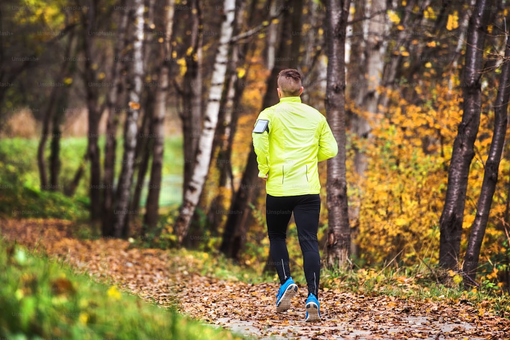 外を走る黄色いジャケットを着たスマートフォンを持った若いアスリート。色とりどりの晴れた秋の自然の中でクロスカントリーランニングのためのトレイルランナートレーニング。