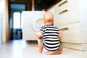 Lindo bebé junto a la lavadora en la cocina. Cesta de la ropa sucia en el suelo. Vista trasera.