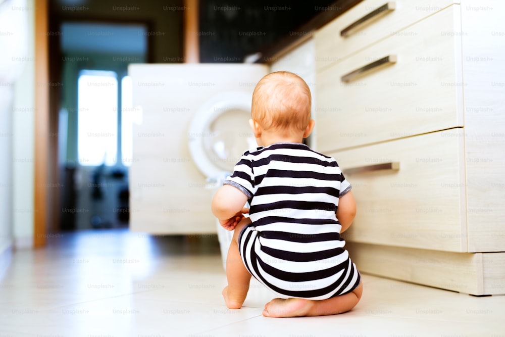 Süßer kleiner Junge an der Waschmaschine in der Küche. Wäschekorb auf dem Boden. Rückansicht.