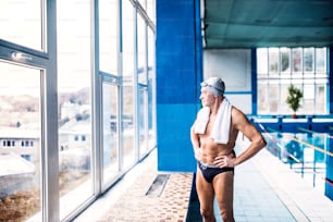 Senior man standing by the indoor swimming pool, arms on hips. Active pensioner enjoying sport.