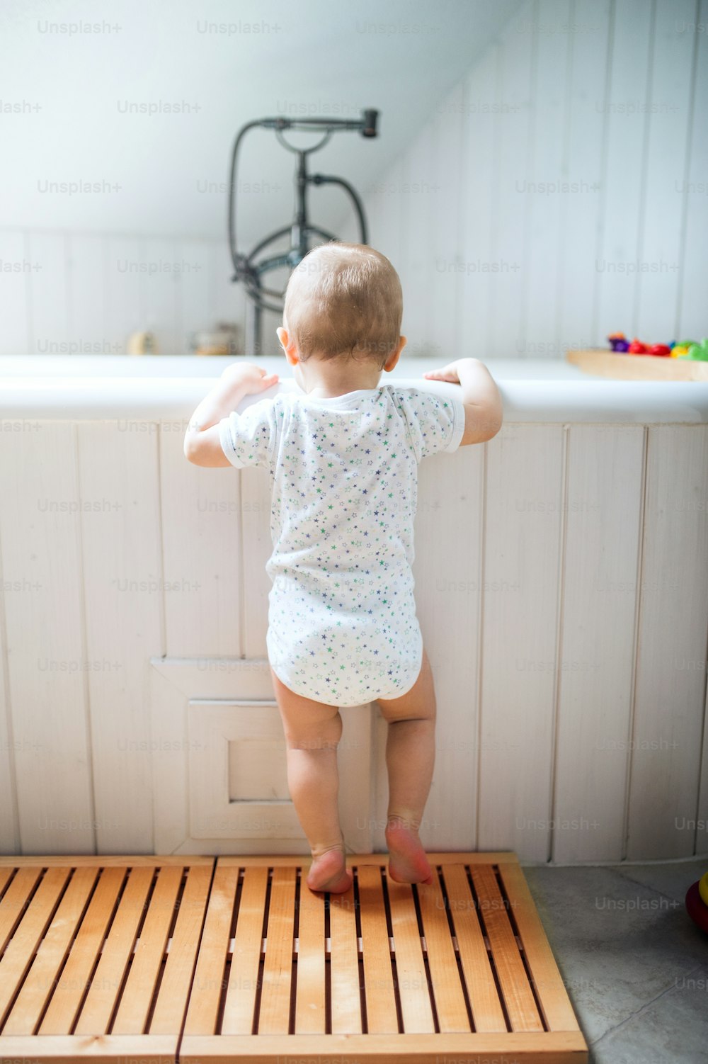 A toddler child standing at the bath in the bathroom at home. Rear view.