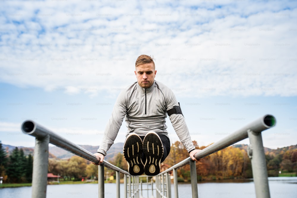 Young man with smartphone in gray jacket exercising outside in autumn nature. Parallel bars L-sit.