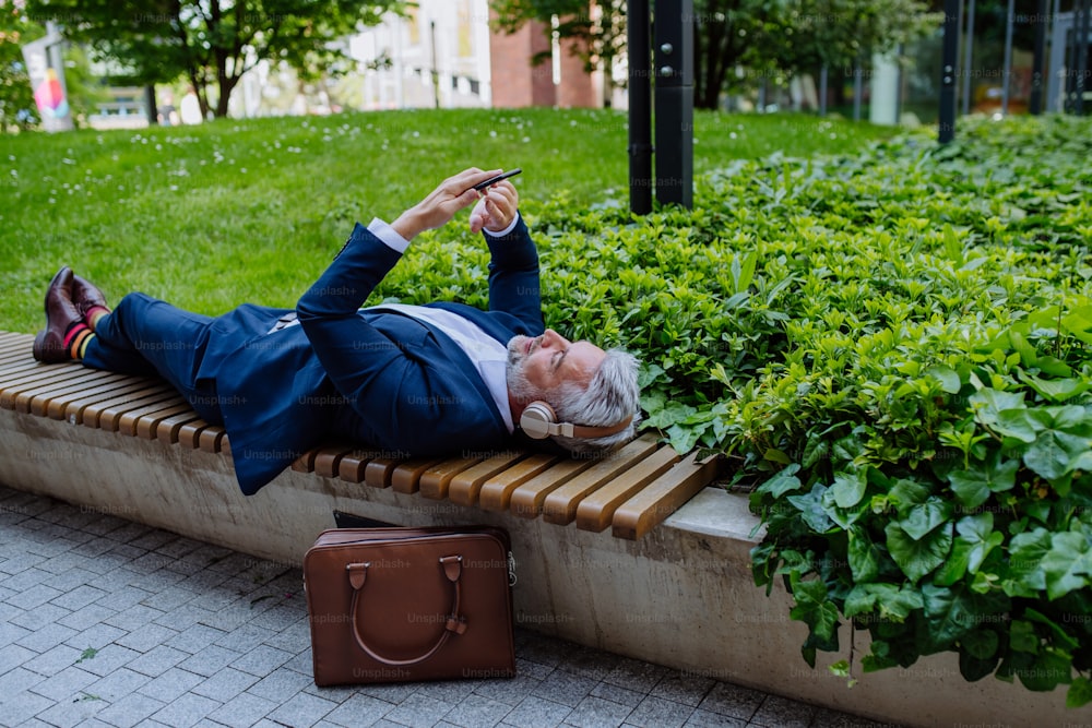 Un homme d’affaires mature appréciant de la musique dans des écouteurs se détendant sur un banc dans un parc de la ville pendant une pause au travail.