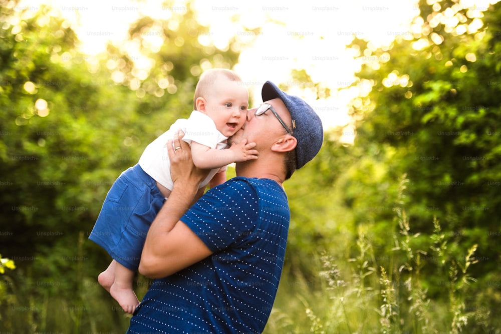 Young father in green summer nature holding his cute baby son in the arms, kissing him on cheek.