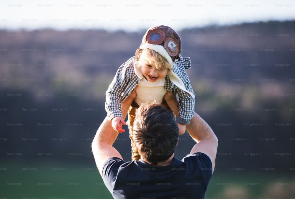 A father lifting his toddler son in the air outside in green sunny spring nature. A boy with pilot goggles and a hat having fun.
