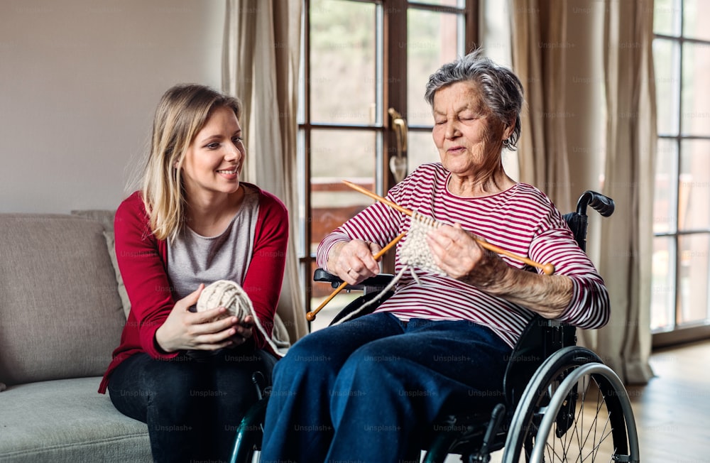 An elderly grandmother in wheelchair with an adult granddaughter sitting on the sofa at home, knitting.