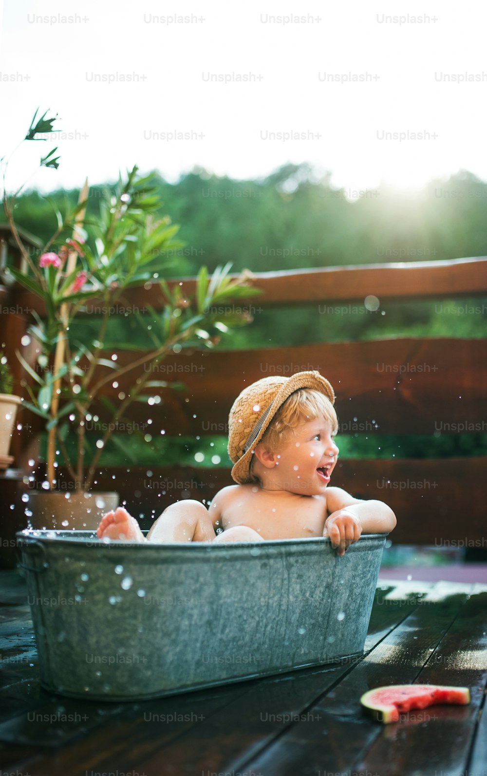 Glücklicher kleiner Junge mit einem Hut in der Badewanne draußen im Garten im Sommer, Wassermelone essend.