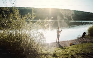 A rear view of a mature man fishing by a lake, holding a rod. A copy space.
