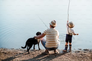A rear view of mature father with a small toddler son and dog outdoors fishing by a river or a lake.