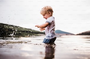 A wet, small toddler boy standing barefoot outdoors in a river in summer, playing with rocks.
