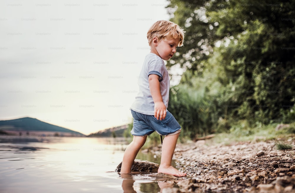 Um menino molhado e pequeno em pé descalço ao ar livre em um rio no verão, brincando.