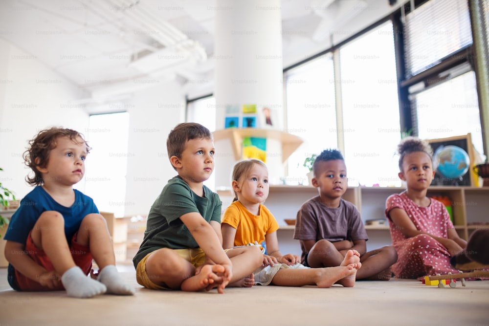 A group of small nursery school children sitting on floor indoors in classroom.