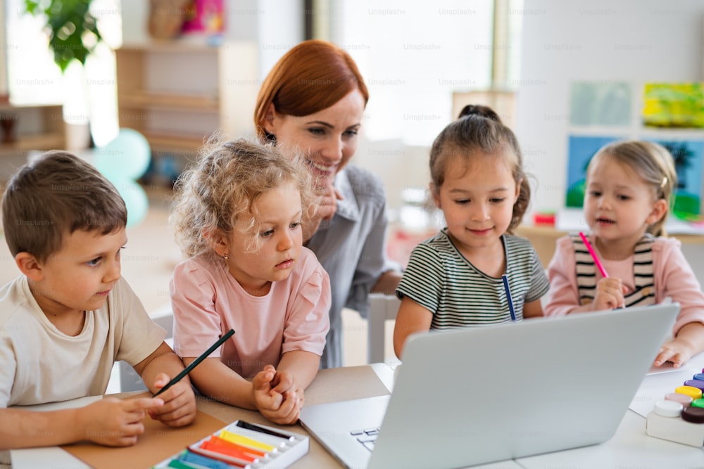 Un groupe de petits enfants de maternelle avec un enseignant à l’étage intérieur de la salle de classe, à l’aide d’un ordinateur portable.