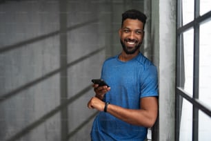 Un joven deportista afroamericano feliz parado en el interior del gimnasio, usando un teléfono inteligente, mirando a la cámara.