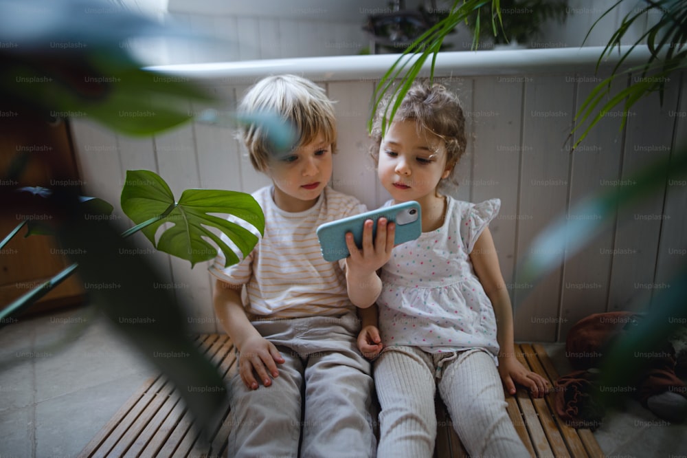 Cute small children sitting on floor indoors in bathroom, using smartphone.