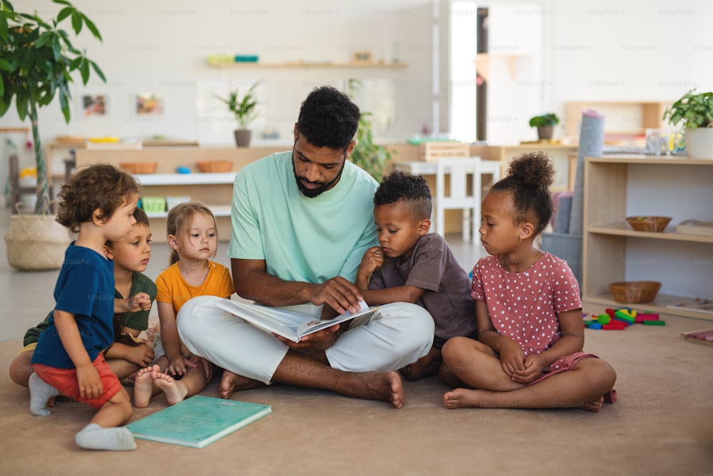Um grupo de pequenas crianças da creche com um professor sentado no chão dentro de casa na sala de aula, lendo um livro de conto de fadas.