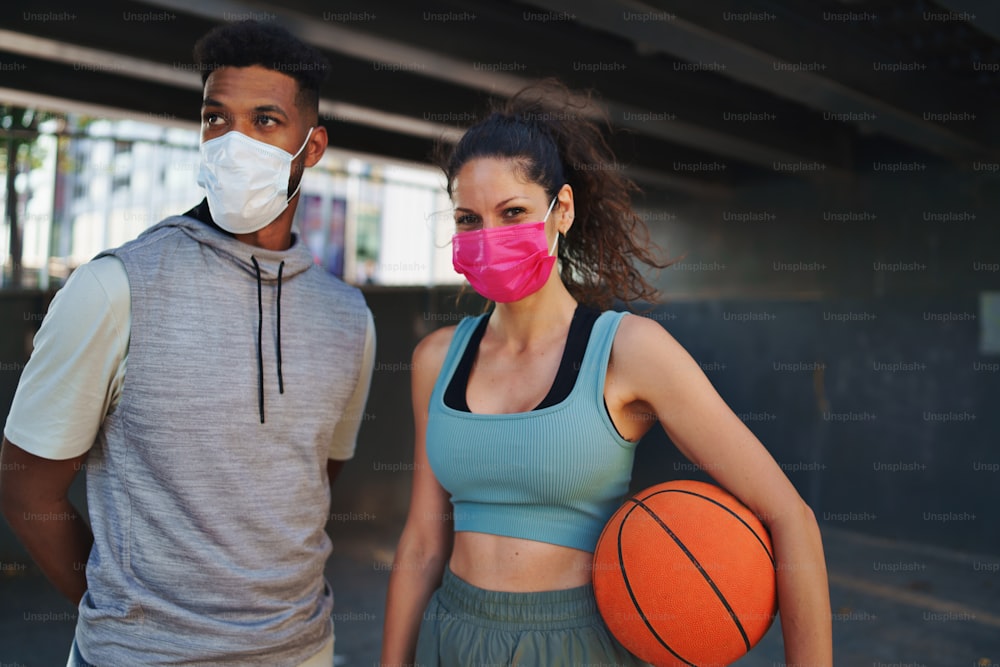 A man and woman friends with basketball doing exercise outdoors in city, looking at camera. Coronavirus concept.