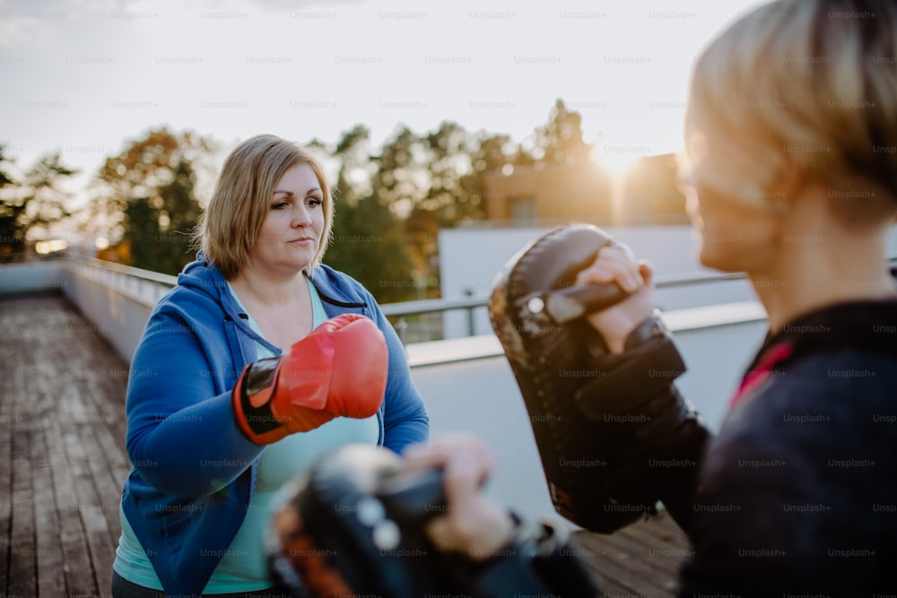 An overweight woman training boxing with personal trainer outdoors on terrace.