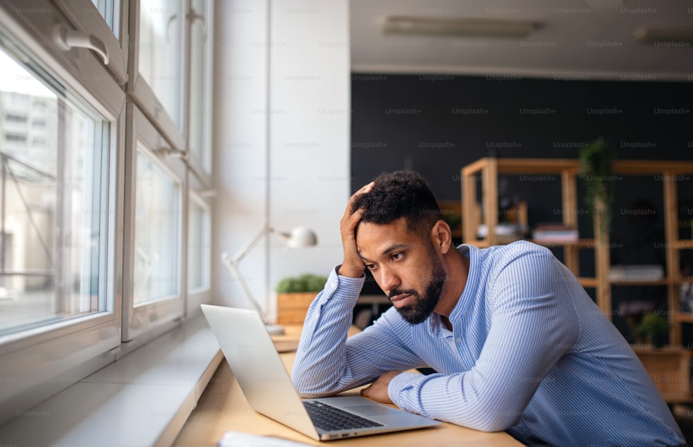 A depressed young African-american teacher using laptop indoors in staffroom.