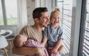 A happy young man taking care of his newborn baby and little daughter indoors at home, looking at camera. Paternity leave.