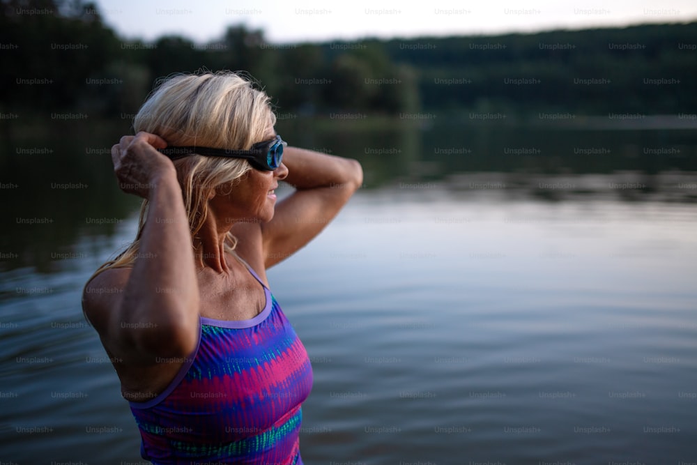 Un portrait d’une nageuse senior active debout et mettant des lunettes à l’extérieur dans un lac.