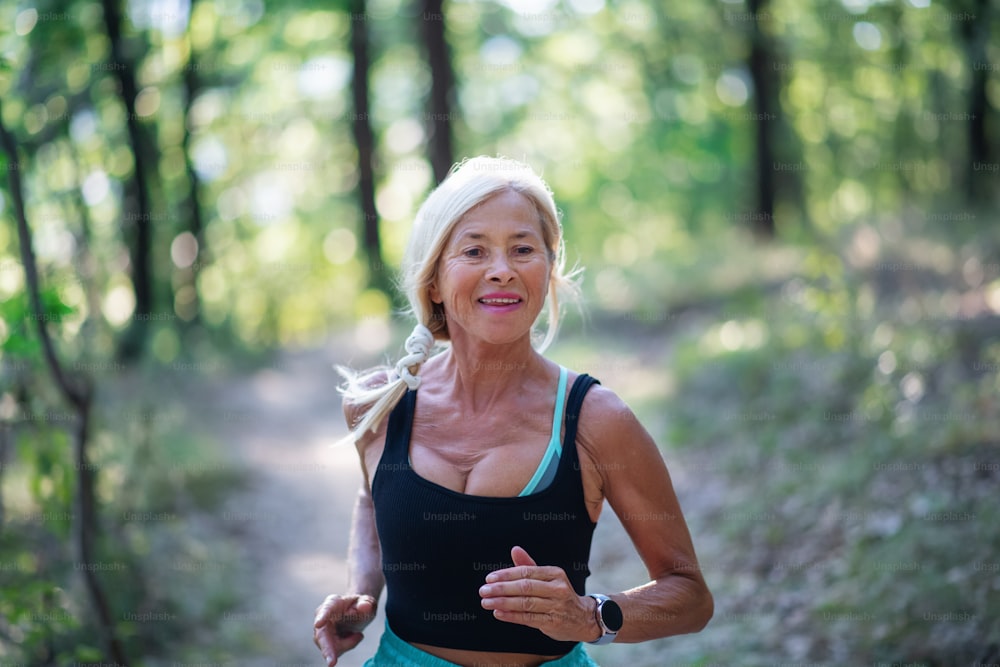 A happy active senior woman jogging outdoors in forest, waist up.