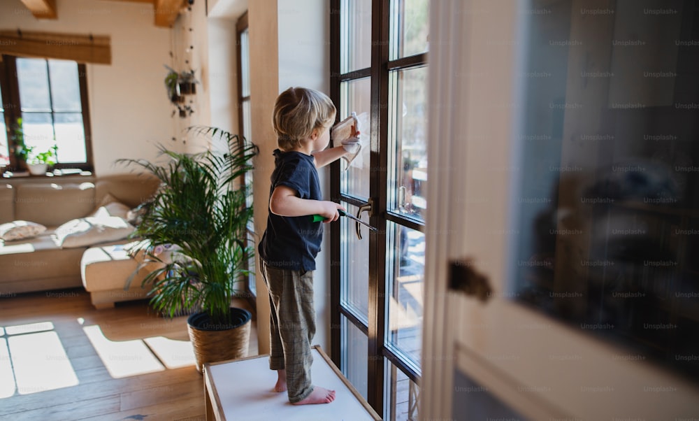 Vista lateral de un niño pequeño limpiando ventanas en el interior de la casa, concepto de tareas diarias.