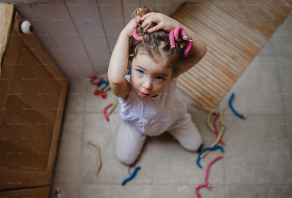 A high angle view of small girl applying hair curlers indoors at home, looking at camera.
