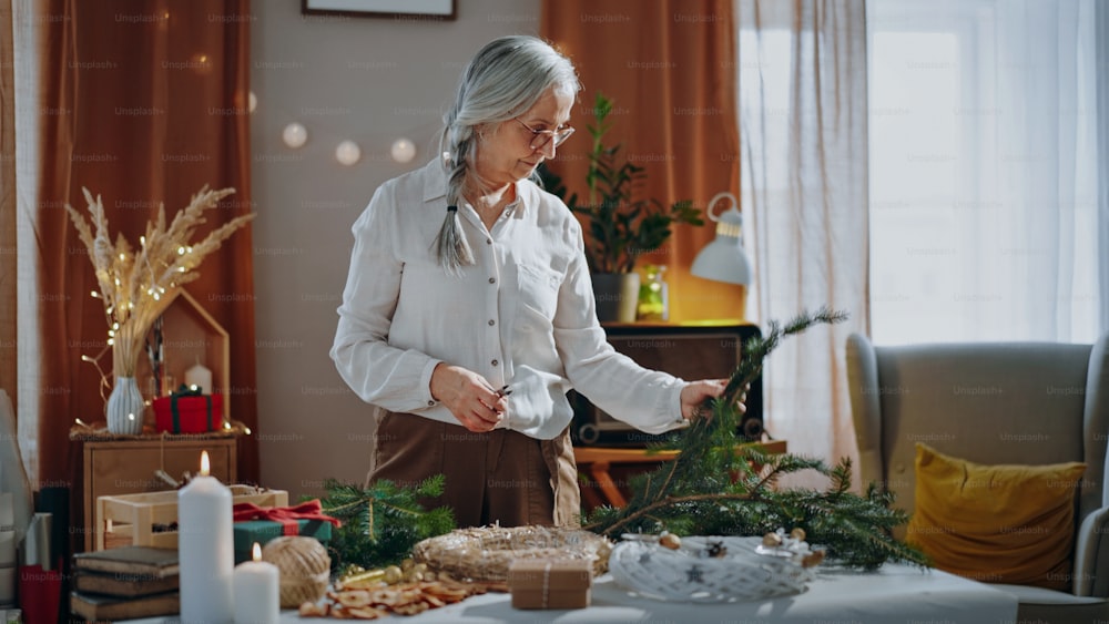 Mulher sênior cortando um ramo para fazer a coroa de Natal dentro de casa em casa