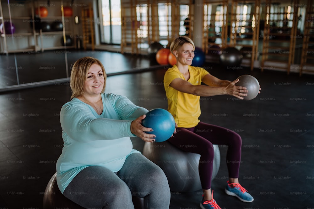 Una mujer feliz con sobrepeso sentada en la pelota fintess y haciendo ejercicio con el entrenador personal en el gimnasio