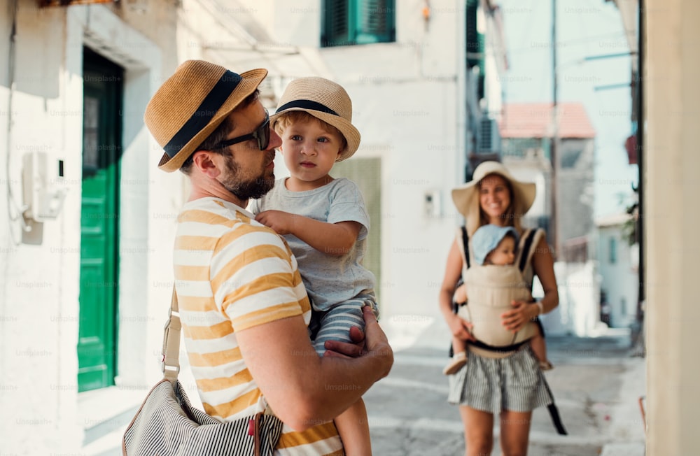 A family with two toddler children walking in town on summer holiday. A father and mother with son and daughter in baby carrier on a narrow street.