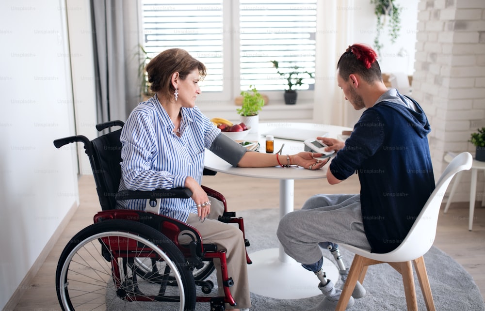 Two disabled people in wheelchair sitting at the table indoors at home, measuring blood pressure.