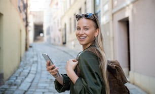 Porträt einer jungen Frau, die im Urlaub in der Stadt mit dem Smartphone in die Kamera schaut.