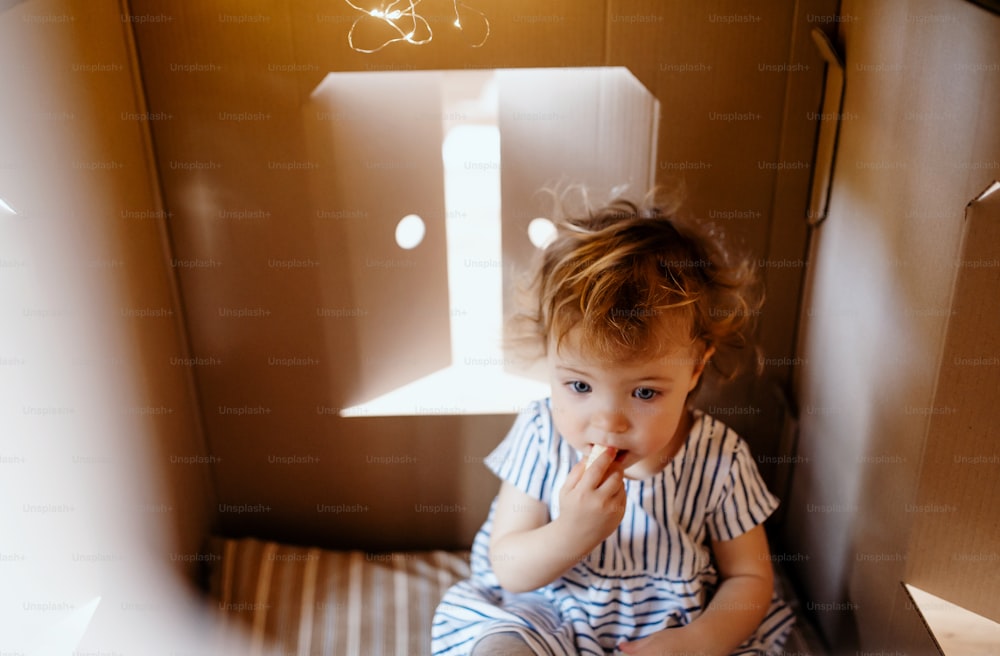 Una niña pequeña jugando en el interior de una casa de cartón en casa, comiendo bocadillos.