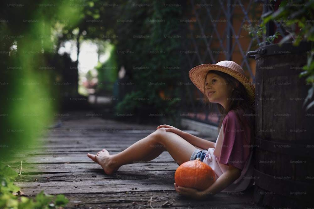 Ein kleines Mädchen mit Hut sitzt und hält Bio-Kürbis draußen auf dem Bauernhof.