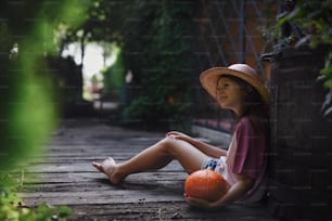 Ein kleines Mädchen mit Hut sitzt und hält Bio-Kürbis draußen auf dem Bauernhof.