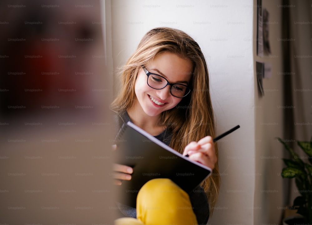 A young happy college female student with an exercise book sitting on window sill at home, studying.