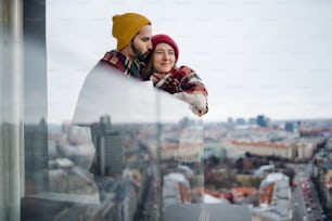 Una giovane coppia avvolta in una coperta in piedi e abbracciata all'aperto sul balcone, ripresa attraverso il vetro.