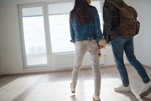 A rear view of unrecognizable young couple owners holding hands in new flat, moving in.