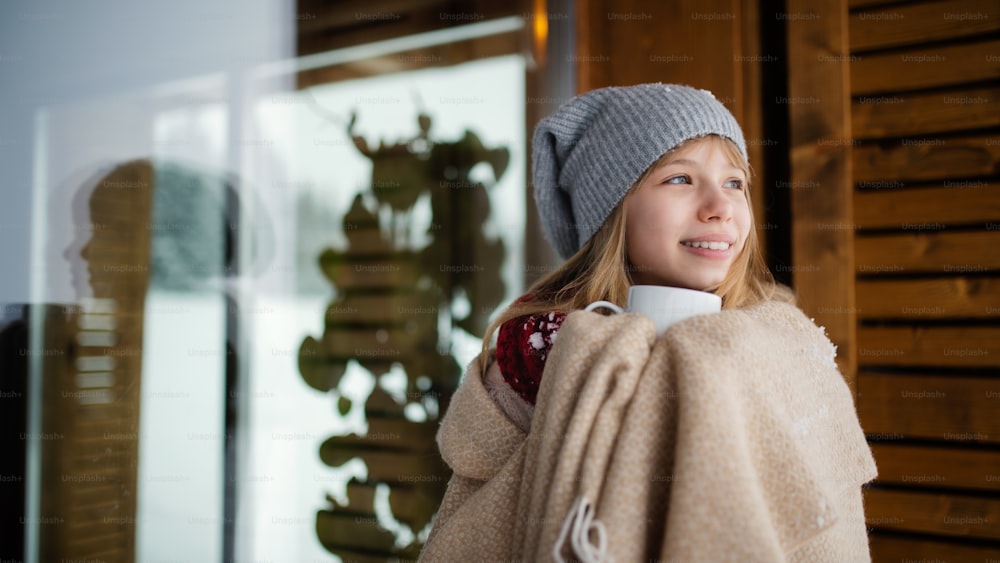 Porträt eines Preteen-Mädchens in Decke gehüllt mit Hoit-Drink im Freien vor einem Holzhaus in der winterlichen Natur.