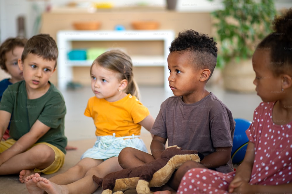 Un grupo de pequeños niños de guardería sentados en el suelo en el interior del aula, escuchando al maestro.