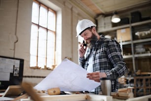 Low angle view of a mature male carpenter looking at blueprints plans in carpentery workshop. Small business concept.